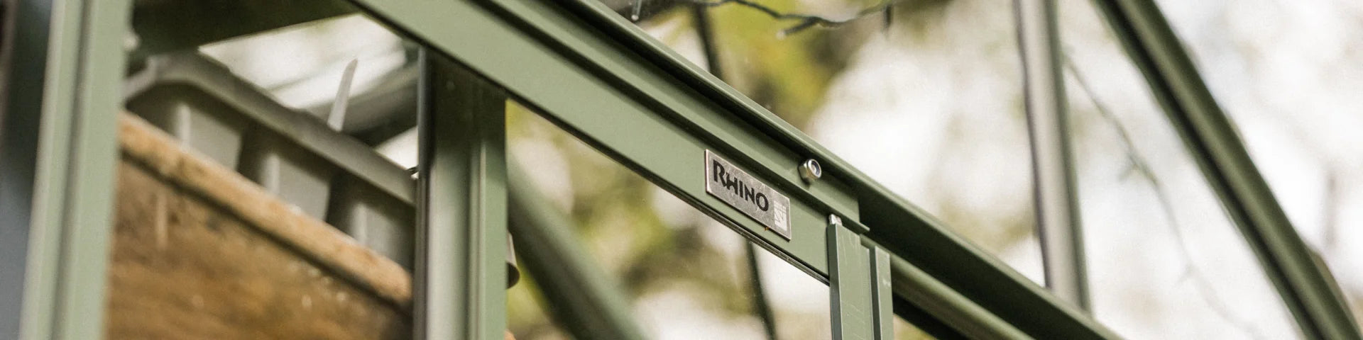 A green metal frame supports a structured Rhino Greenhouse with a visible Rhino label, surrounded by blurred outdoor foliage.