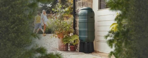 A green water butt stands by a white building, surrounded by potted plants. In the background, a person and child walk along a gravel path, framed by lush greenery.
